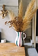 A white ceramic pitcher with vertical pink, green, and black stripes, filled with dried pampas grass and brown botanicals, set on a wooden cutting board on a kitchen counter next to a bottle of olive oil.