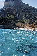 A scenic coastal photograph showing bright turquoise water in the foreground and a steep rocky cliff covered with green vegetation rising behind it under a pale blue sky.