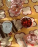 A styled tabletop scene with a glass pitcher of pink drink, a bowl of red cherries, two glasses with ice, and a lime wedge on a patterned yellow tablecloth.