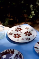 A white ceramic plate with a dark brown floral pattern, set on a blue tablecloth outdoors with a glass cup nearby.
