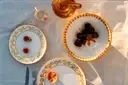 Three white ceramic plates with gold scrollwork borders are arranged on a light beige linen tablecloth, styled with a small orange cup, a glass teapot, and food items under warm sunlight.
