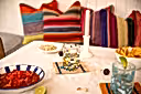 A styled tabletop scene with colorful striped pillows in the background, a lit pink taper candle in a white holder, a glass tumbler with multicolored stones, bowls of salsa and chips, and a blue-tinted drink in the foreground on a white tablecloth.