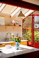 A bright kitchen interior with a marble countertop in the foreground, a blue-and-white striped vase holding colorful flowers, wooden cabinetry, a sink, and a red-framed glass door opening to greenery outside.